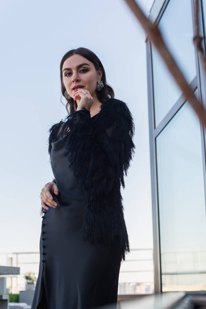 low angle view of young woman in black slip dress and faux fur jacket on terrace  - Fotó, kép
