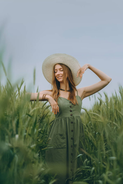 Mujer joven en un sombrero caminando en un campo de trigo, disfruta de la vida y el verano. Campo de trigo. Hermoso paisaje natural bajo la luz del sol. Concepto de estilo de vida saludable. Imagen con ruido, grano de película - Foto, Imagen