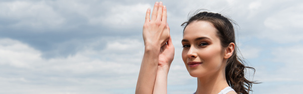 brunette woman with clenched hands looking at camera against blue sky, banner - Фото, зображення