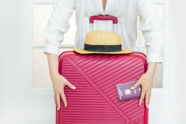 Mujer con maleta rosa sostiene sombrero de verano y pasaporte rumano y listo para viajar. - Foto, imagen