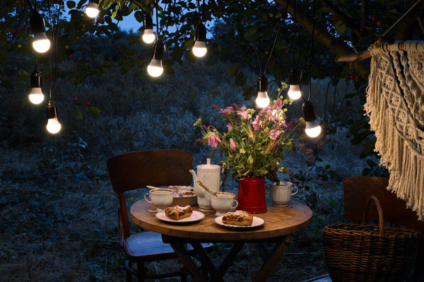 tea party in the evening garden. on the table is a vase of flowers, pie with cherry and cup of tea. garland and festive lightin - Фото, изображение