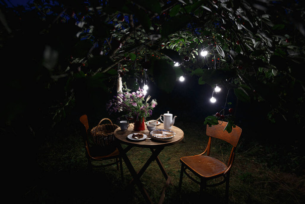 tea party in the evening garden. on the table is a vase of flowers, pie with cherry and cup of tea. garland and festive lightin - Photo, image