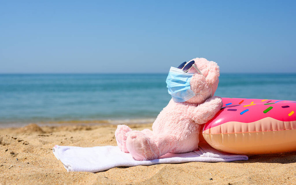 Плюшевый розовый цвет на пляже загорает в медицинской маске и солнцезащитных очках. Концепция рекреации в контексте эпидемии коронавируса. - Фото, изображение