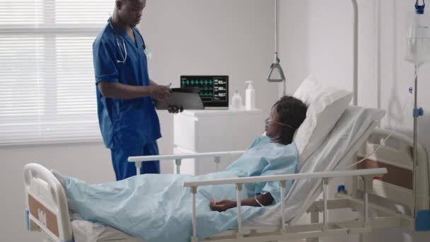 Egy fekete férfi orvos beszél egy fekete női beteggel, aki egy kórházi ágyon fekszik, és egy savellátmányhoz kapcsolódik. A kezelőorvos megbeszéli a kórházban lévő beteggel a kezelést. - Felvétel, videó