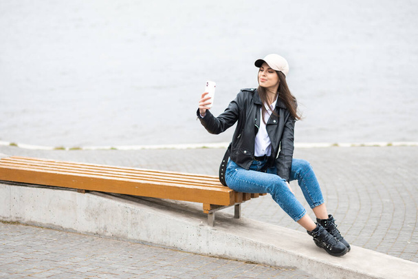 A young pretty girl, dressed in a leather jacket and jeans, spends time alone, sitting on a bench by the river. - Fotografie, Obrázek