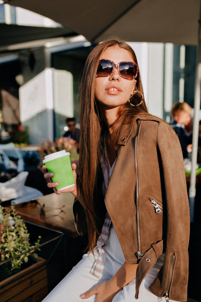 Attraktive, stylische Frau mit langen braunen Haaren, Jacke und Sonnenbrille, die draußen mit Kaffee im Sonnenlicht sitzt. Hochwertiges Foto - Foto, Bild