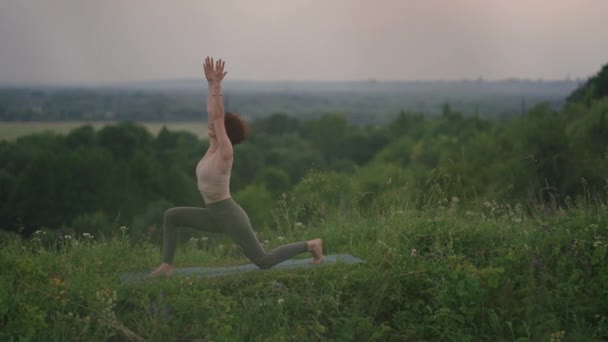 若い女性が川や森の風景を背景に森の山の上に立つヨガの練習をします。身体と精神のバランス、リラクゼーションと瞑想  - 映像、動画