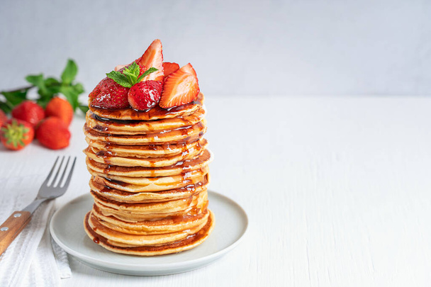 Pile of homemade pancakes or crepes decorated with strawberries, mint leaves and syrup topping served on gray round plate on white wooden table with fork and towel. Horizontal image with copy space - Фото, изображение