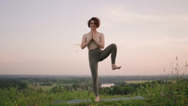 Uma jovem faz exercícios de ioga em pé no topo de uma montanha florestal contra o fundo de um rio e uma paisagem florestal. Equilíbrio de corpo e espírito, relaxamento e meditação  - Filmagem, Vídeo
