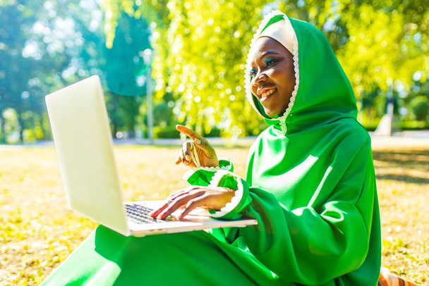 hispanic woman in green muslim hijab with bright make up and nose piercing working by laptop sit on grass outdoors summer park - Foto, Imagen