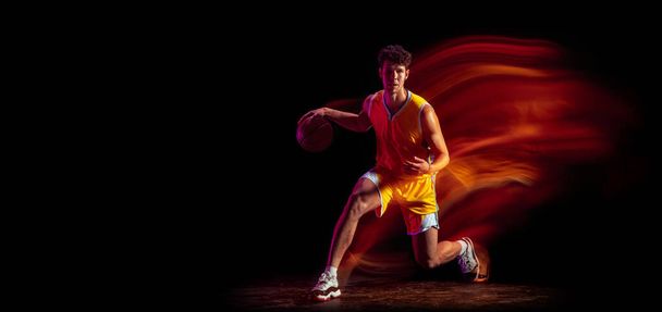 Joven jugador de baloncesto profesional caucásico haciendo ejercicio aislado sobre fondo negro en luces de neón mixtas. - Foto, imagen