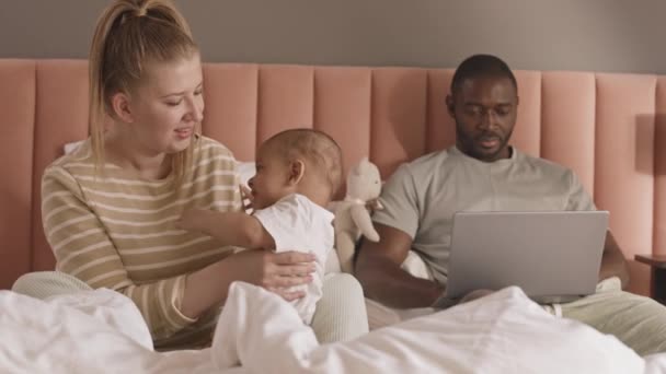 Medio lungo di bionda donna caucasica ammirando il suo bambino felice, seduto sul letto, giovane marito africano che utilizza il computer portatile su sfondo - Filmati, video