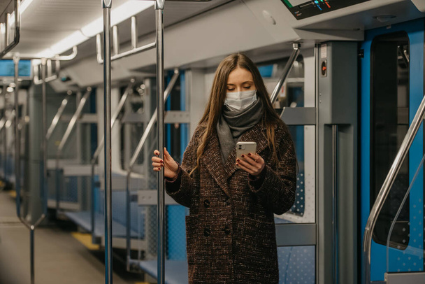 Una mujer con una máscara facial médica para evitar la propagación del coronavirus está de pie y usando un teléfono inteligente en un vagón de metro vacío. Una chica con una máscara quirúrgica está escribiendo noticias en su celular en un tren.. - Foto, Imagen