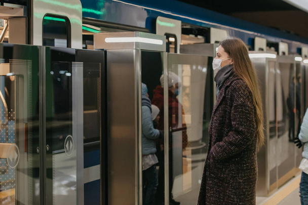 Uma mulher em uma máscara facial médica para evitar a propagação do coronavírus está entrando no moderno carro do metrô. Uma menina em uma máscara cirúrgica está mantendo a distância social em uma estação de metro. - Foto, Imagem