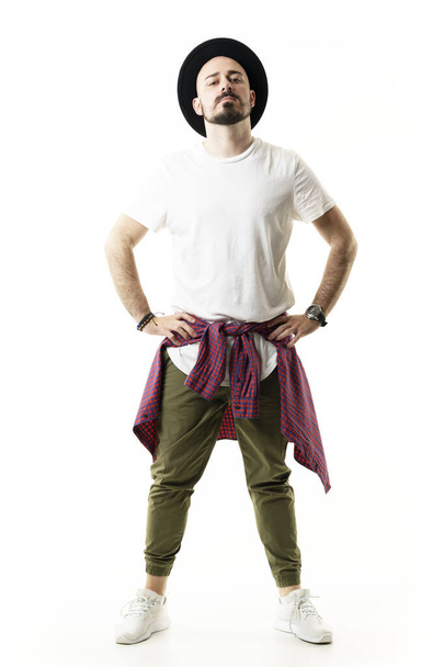 Zelfverzekerde eigenwijze hipster met hoed in chino broek poserend met handen op de heupen. Full body portret geïsoleerd op witte achtergrond - Foto, afbeelding