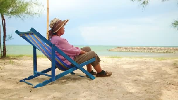 Μια ηλικιωμένη Ασιάτισσα με καπέλο κάθεται σε μια ξαπλώστρα δίπλα στην παραλία. Η έννοια του ηλικιωμένου ταξιδιωτικού χαρακτήρα κατά την ηλικία συνταξιοδότησης. Αντιγραφή χώρου - Πλάνα, βίντεο