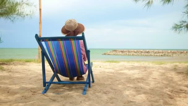 Een oudere Aziatische vrouw met een hoed zit op een ligstoel aan het strand. Het concept van de bejaarden reizen de natuur tijdens de pensioengerechtigde leeftijd. Kopieer ruimte. - Video