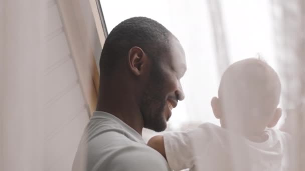 Ώμο-up backlight του νεαρός Αφρικανός κρατώντας μικτή φυλή μωρό του, μιλάμε για αυτόν και χαμογελώντας πίσω από κουρτίνες από το παράθυρο - Πλάνα, βίντεο