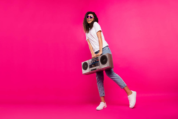 Full body photo of pretty millennial lady hold boom box wear spectacles t-shirt jeans sneakers isolated on pink background - 写真・画像