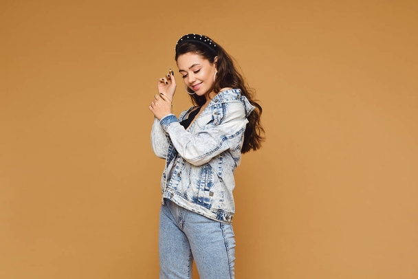 Caucasian young woman with wavy hair in denim clothes, isolated on beige. Model girl in a modish outfit at the beige background - 写真・画像