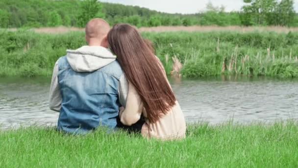 słodka para siedzi nad rzeką na zielonej trawie. atrakcyjna brunetka i łysy mężczyzna na świeżym powietrzu. Prawdziwa miłość, szczęśliwa rodzina, romans, udane małżeństwo. zwolniony ruch - Materiał filmowy, wideo
