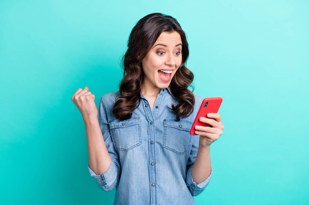 Photo of pretty sweet young lady dressed denim shirt smiling looking modern gadget rising fist isolated teal color background - 写真・画像
