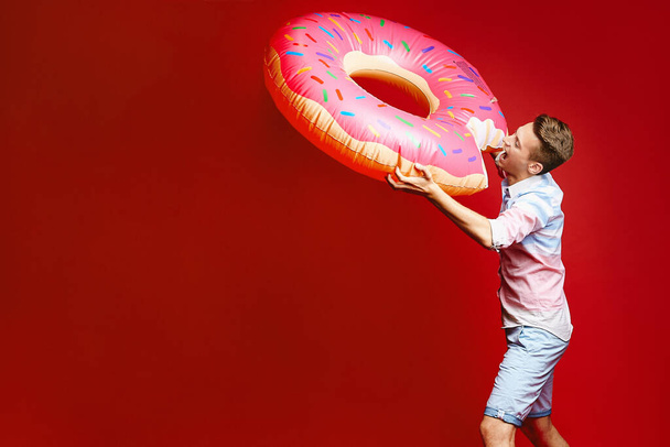 A young stylish blond man in a sleeveless shirt and shorts posing with an inflatable donut, isolated at red background. A young handsome guy in a summer outfit posing over red background - Foto, Bild