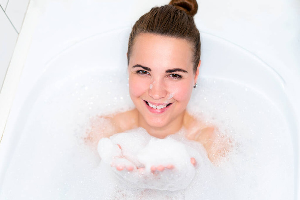Beauty portrait of beautiful happy positive girl, young cheerful woman is relaxing, lying in bathroom, taking a bath with bubbles, smiling and looking at camera, washing her body with foam in bathtub - Valokuva, kuva