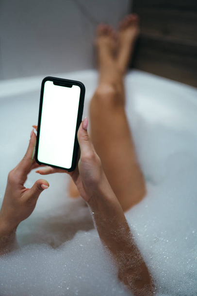 Photo of female legs lying in a foam bath and hands holding a phone with a light screen. Space for text - Zdjęcie, obraz