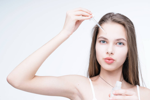 A beautiful girl with long hair applies a serum to her face while holding a pipette in her hand - Foto, immagini