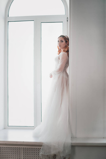 een full-length portret van een mooie bruid met rood haar in een witte transparante jurk staat in de boog van het raam op een bruiloft ochtend - Foto, afbeelding