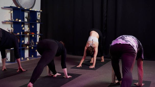 Drie meisjes en twee jongens tijdens yogasessie in sportstudio met zwarte gordijnen - Foto, afbeelding