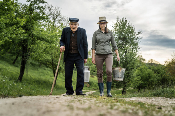 Vorderansicht des erwachsenen Rentners mit seiner Enkelin erwachsene kaukasische Frau zu Fuß auf Feldweg im Freien halten Milcheimer - echte Menschen Mehrgenerationenlandwirtschaft Konzept - Foto, Bild