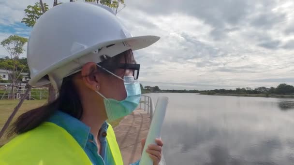 白いヘルメットと保護マスクを身に着けている黄色の制服の女性エンジニアは青写真を保持し、電気を生成するためにダム建設現場を離れて見て. - 映像、動画