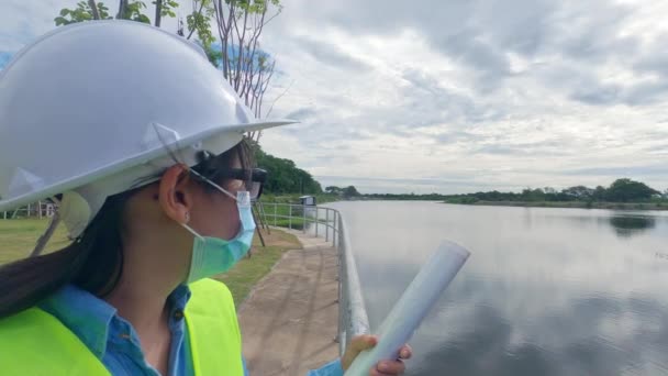 Female engineer in a white helmet and yellow uniform wearing a protective mask holds a blueprint and looking away at the dam construction site to generate electricity. - Séquence, vidéo