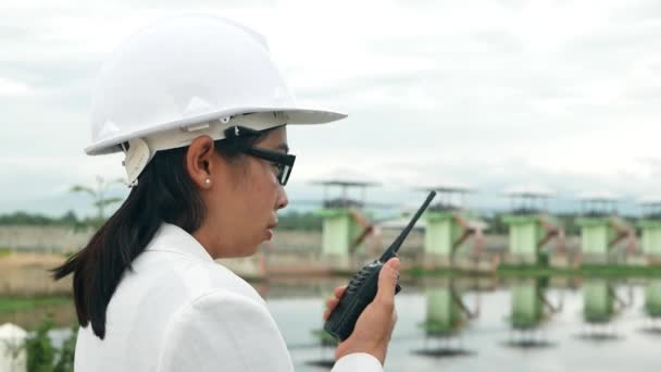 Female engineer in a white helmet using radio communication with worker at the dam construction site to generate electricity. - Séquence, vidéo