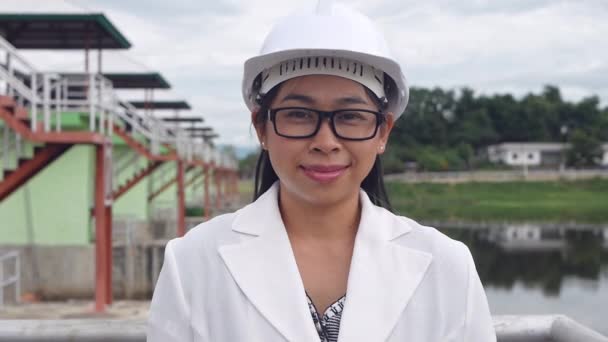 Γυναίκα μηχανικός σε ένα λευκό κράνος κοιτάζει και χαμογελά σε μια κάμερα σε ένα εργοτάξιο φράγμα για την παραγωγή ηλεκτρικής ενέργειας. - Πλάνα, βίντεο