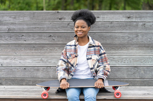 Feliz sonriente mujer africana con longboard skate en ropa casual y ganancias de moda sentarse en el banco - Foto, Imagen