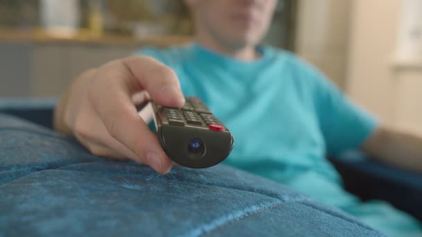 Primer plano de una mano de hombre con un mando a distancia de televisión sentado en casa en el sofá - Imágenes, Vídeo