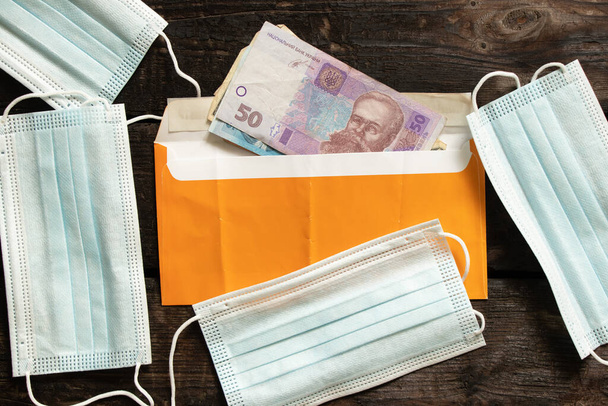 медицинская маска лежит в конверте с украинскими деньгами на деревянном столе, деньгами и медицинской маской - Фото, изображение