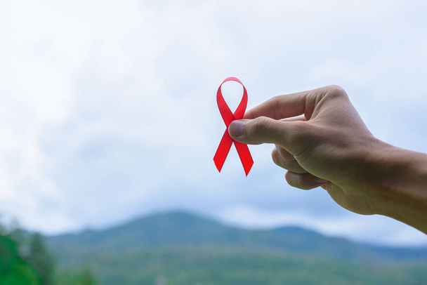 Ruban de sensibilisation à la Journée mondiale du sida, mains féminines tenant un ruban rouge sur fond de nature VIH, concept de la Journée mondiale du sida. - Photo, image