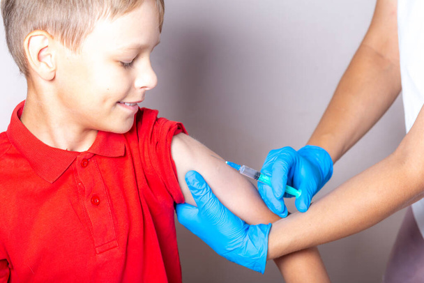 A nurse in a white cap, disposable mask and blue gloves injects the vaccine through a syringe into the shoulder of a brave boy in a red T-shirt. - 写真・画像