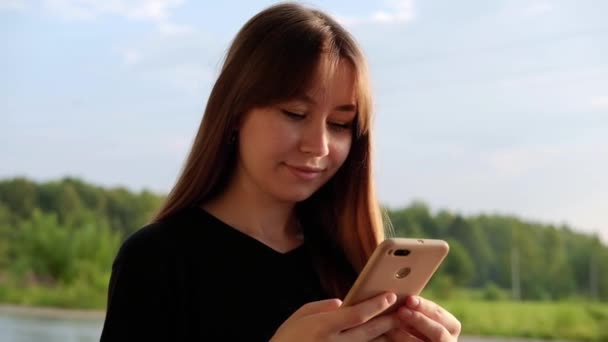 Mujer joven sonriente en camiseta negra usando un teléfono inteligente, desplazándose por el campo. Usando dispositivo. Chica mirando hacia abajo y mensajes de texto en el teléfono inteligente. Chat, mensajería, lectura. - Metraje, vídeo