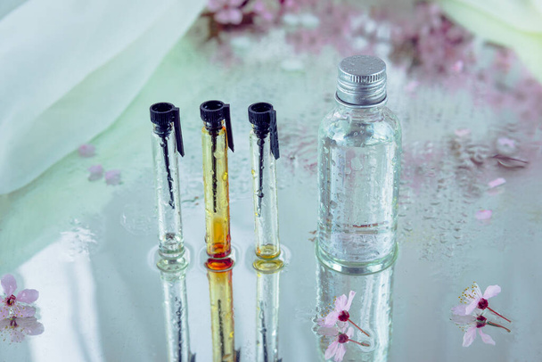 Zestaw trzech testerów szkła i butelka pokryta kroplami wody i umieszczona na mokrym lustrze wraz z niewyraźnym różowym kwiatem. Koncepcja olejków eterycznych i ekstraktów ziołowych. - Zdjęcie, obraz