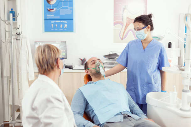 Ορθοδοντικός βοηθός βάζοντας μάσκα οξυγόνου στον ασθενή αναλύοντας τον πόνο των δοντιών - Φωτογραφία, εικόνα