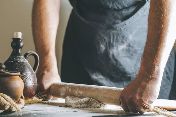 Männliche Hände rollen Teig und Mehl mit Nudelholz neben Tontopf und Ölflasche auf dunklem Tisch beim Kochen - Foto, Bild
