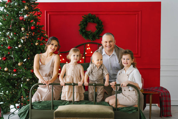 Stylowa rodzina z trojgiem dzieci siedzi na łóżku obok choinki. Przytulne święta Bożego Narodzenia - Zdjęcie, obraz