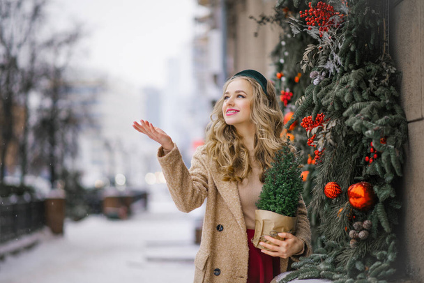 Una guapa rubia sostiene un árbol de Navidad en maceta en sus manos, que compró en una tienda, y camina por la ciudad en un día de Navidad de invierno. Le ofrecí mi palma a la nieve atacante. - Foto, imagen