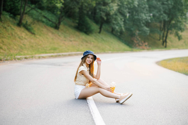 A happy girl in white shorts and a yellow blouse holds a glass of juice with a straw in her hands and sits on the road near the forest - Photo, Image