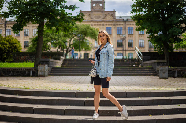 Young beautiful blonde woman in a denim jacket and tight sports shorts stands on the stairs and looks straight - Фото, изображение